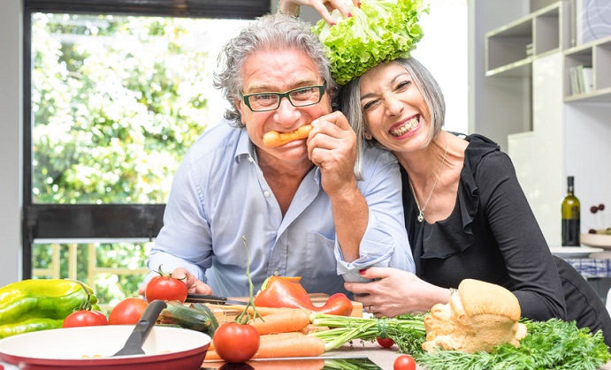 senior couple having fun in kitchen with healthy food - retired people cooking meal at home with man and woman preparing lunch with bio vegetables - happy elderly concept with mature funny pensioner