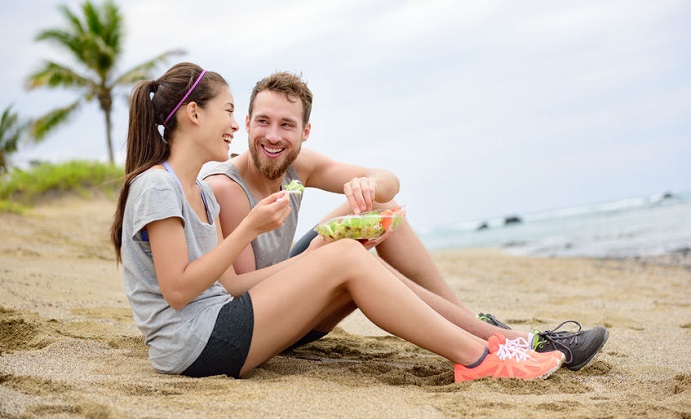 salad - healthy fitness woman and man couple laughing eating food lunch sitting on beach after workout. mixed race asian caucasian female model and male models in sportswear.